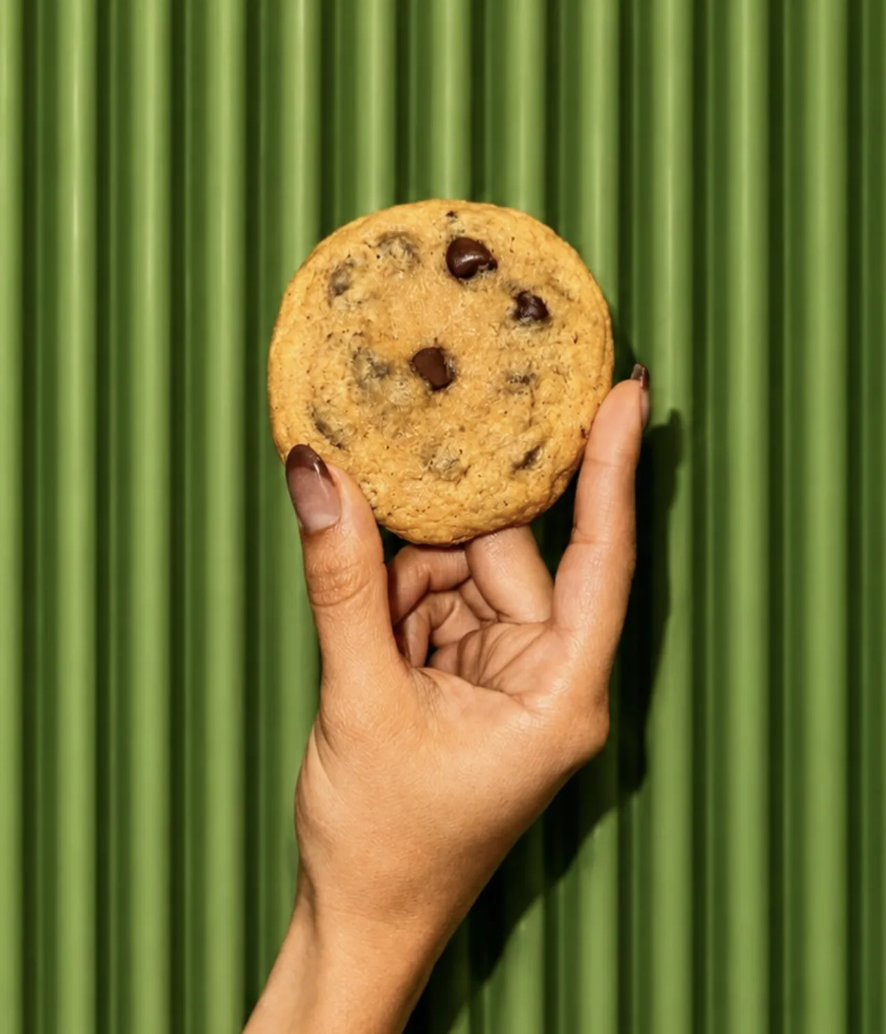 Hand holding a chocolate chip cookie against a green ribbed wall