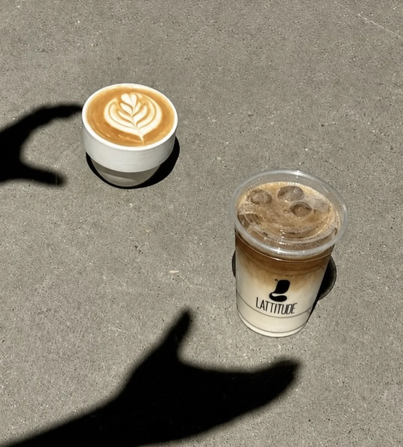 Overhead shot of a latte with leaf art and an iced coffee to go, a hand's shadow crossing the pavement